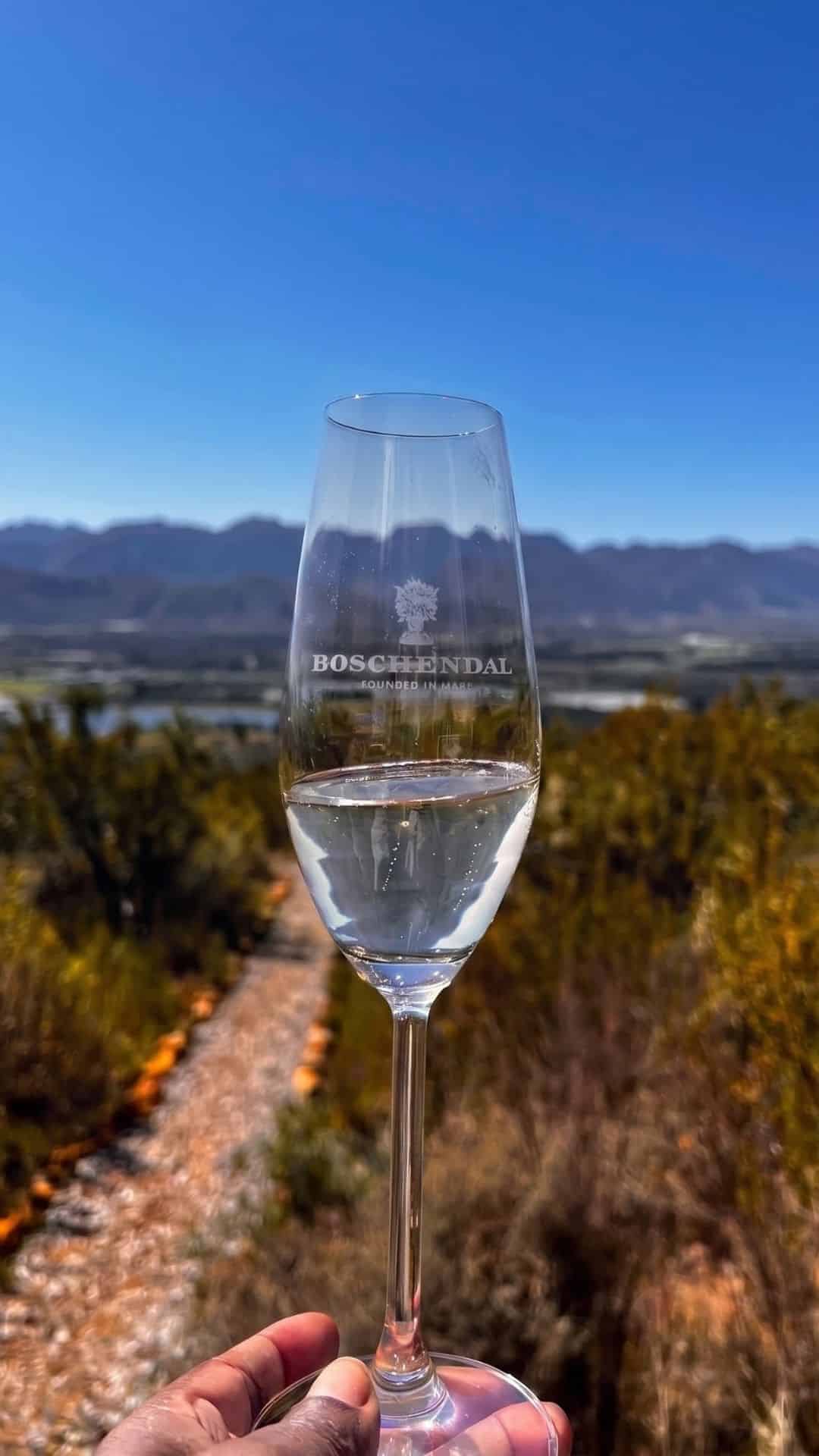 Boschendal-etched wine glass held up over a vineyard path at a Stellenbosch wine farm